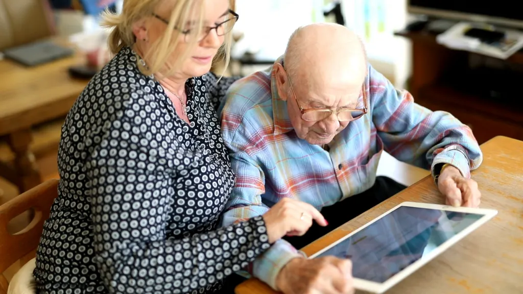 Eine Pflegekraft aus Polen und der alte Mann den sie betreut betrachten eine Webseite auf einem Ipad